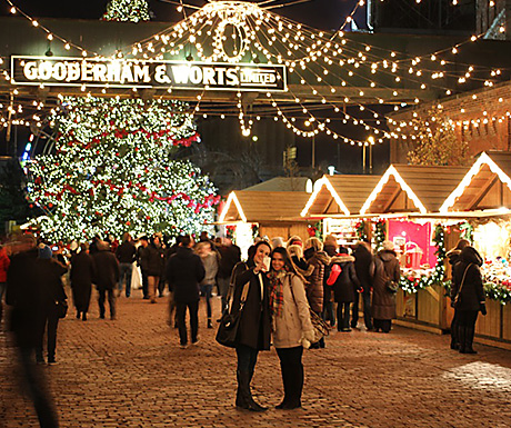 two women take a selfie in front of a brightly lit Christmas market in Toronto