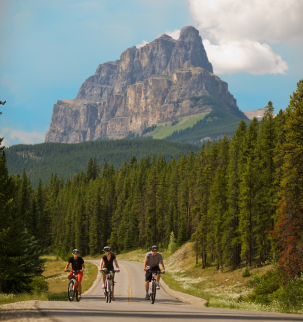 three mountain bikers cycle up a road away from a giant mountain peak