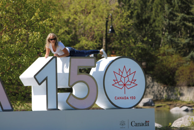 woman lies across the Canada 150 sign with trees in the background