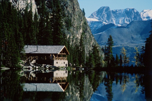 log cabin and mountains behind are reflected in the waters of a lake