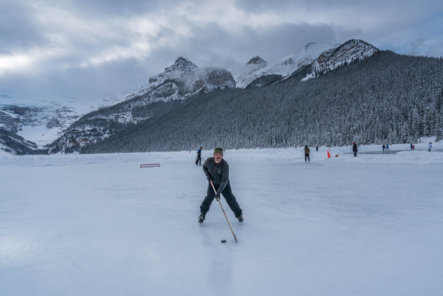 man plays ice hockey on frozen lake with the Rocky Mountains behind him