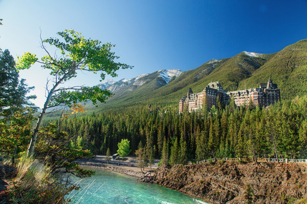 a grand hotel sits nestled above a flowing turquoise river in the mountains