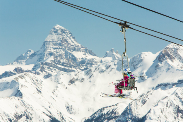 two skiers in pink coats ascend in a chair lift with a huge mountain peak in the background