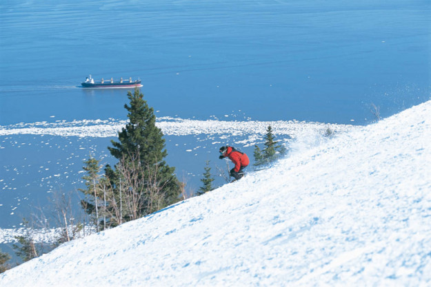 a skier in a red jacket descends a slope with a cargo ship out at sea in the background