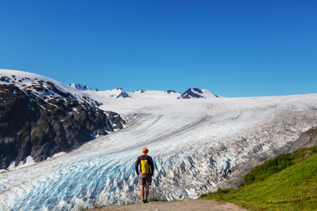 a man with a yellow backpack stands at the edge of a glacier with grass to his right