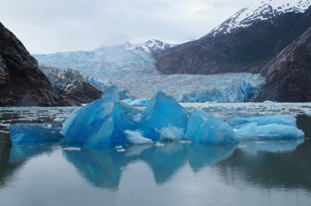 a bright blue iceberg sits in grey water