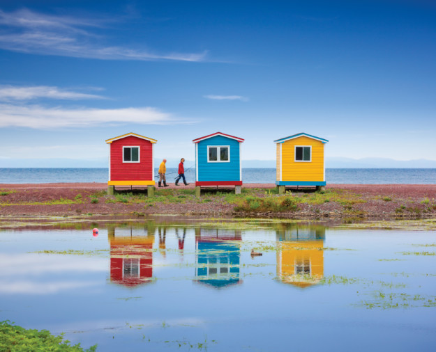 a couple walk behind multi-coloured fishing huts on the water