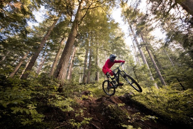 a mountain biker in a red jacket takes a jump in the forest