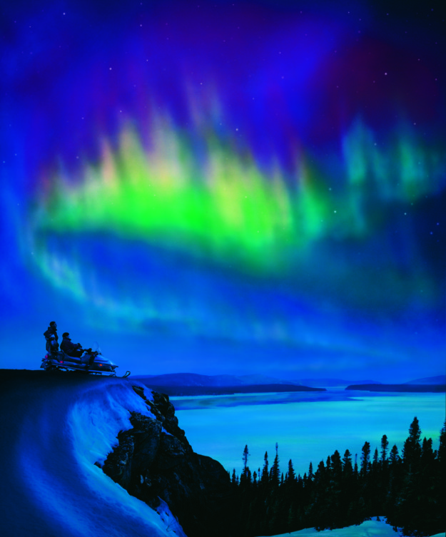 two people on a snowmobile on the edge of a cliff watch the northern lights in the sky