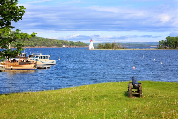 a cannon sits on a grassy field next to inlet of water with boats and a lighthouse in the background