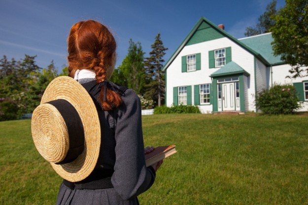 a girl in a grey dress with red hair and and straw boater hat holds a book in one hand and looks at the green and white wooden plank Anne of Green Gables house