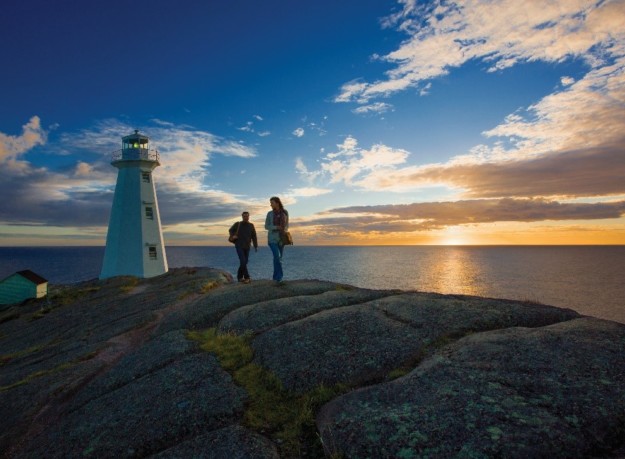 couple hike along rocks with a white lighthouse and the rising sun in the background