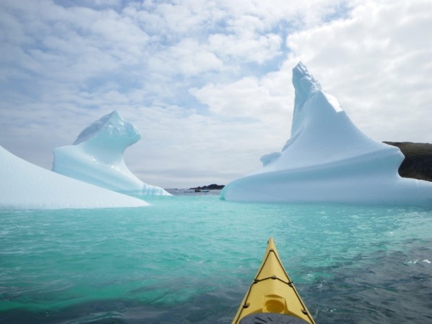 the front of a yellow kayak drifts on turquoise water towards three icebergs