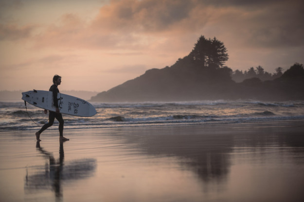 a surfer carries their board walking along the beach in the early morning