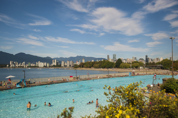people enjoy a large swimming pool on a sunny day with the Vancouver skyline in the background