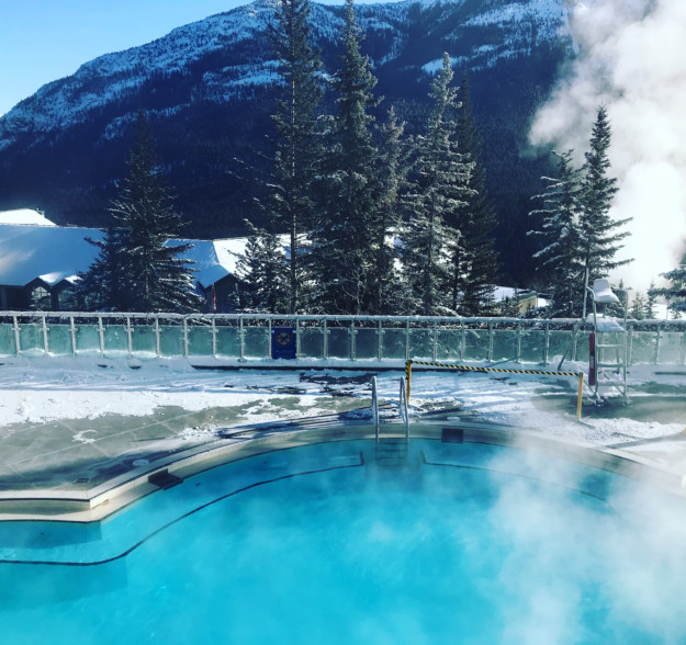 steam rises off a heated pool with snowy mountain forest in the background