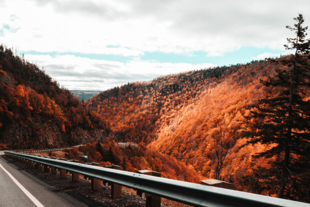 A scenic roadway in Nova Scotia bending through a forested landscape with vibrant orange and red autumn foliage under a cloudy sky.