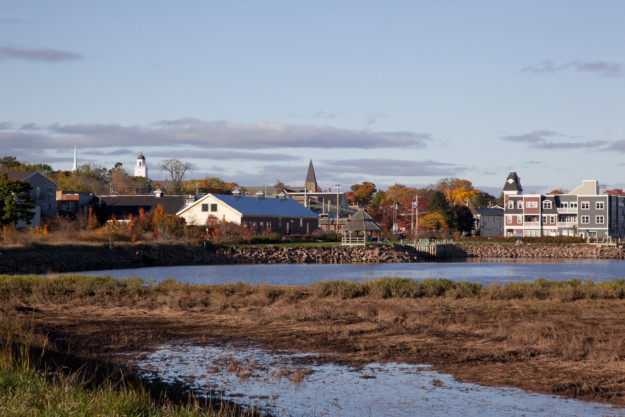 A picturesque view of a small town in Nova Scotia by a river, with colorful autumn trees and traditional houses. A clear sky and reflective water enhance the tranquil scenery.