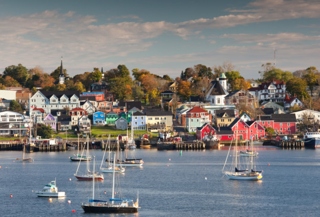 A picturesque coastal town in Nova Scotia with colorful buildings along the waterfront, dotted with moored sailboats on a sunny day, under a clear blue sky.