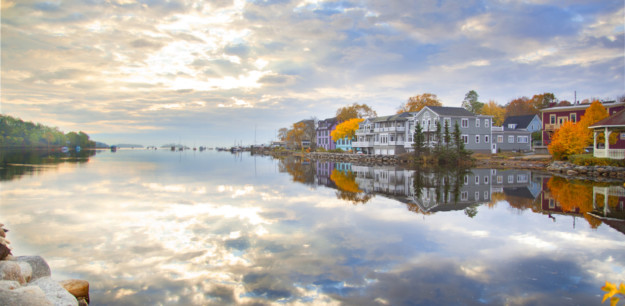 A serene waterfront scene in Nova Scotia, with buildings reflecting in calm waters, under a sky dotted with clouds during sunrise or sunset, and autumn-colored trees visible.