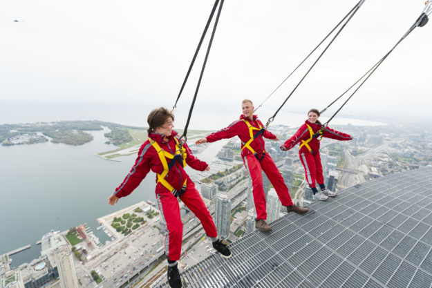 Three people in red jumpsuits and harnesses walk on a metal grate platform high above a cityscape with lake views in Ontario, holding onto safety cables.