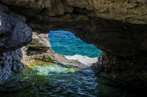 A view through a rocky cave opening in Ontario, leading to the sea, with turquoise waters visible and sunlight illuminating the surrounding rocks.