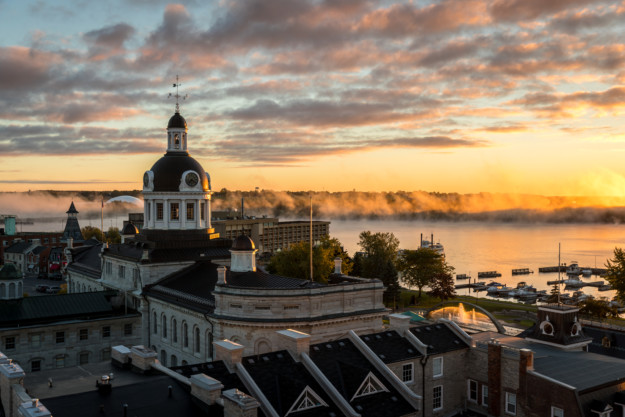 Sunrise view over a historic building with a domed rooftop in Ontario, overlooking a river shrouded in mist, with boats docked beside it and trees in the distance under a glowing sky.
