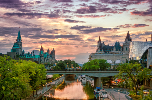A scenic view of a cityscape at sunset in Ontario featuring historic buildings with spires, a modern glass structure, and a river running through, flanked by green trees.