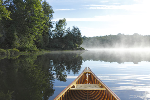 The front of a wooden canoe on a misty lake in Ontario, with lush green trees lining the shore under a clear sky.