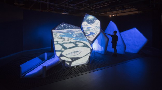A person interacts with a magical art installation featuring luminous abstract projections on pyramid-like shapes in a dimly lit room.