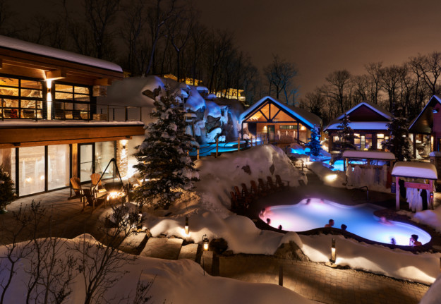 A luxurious winter resort at night in Canada's capital, featuring illuminated buildings with large windows, a heated outdoor pool surrounded by snow, and trees gently dusted with snow under a serene, dark sky.