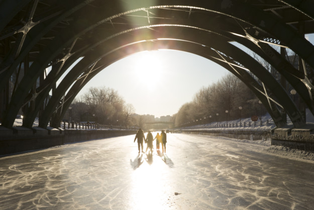 Group of people walking on a frozen river under a large arched bridge in Canada's capital, bathed in sunlight. The low sun creates long shadows on the icy surface.