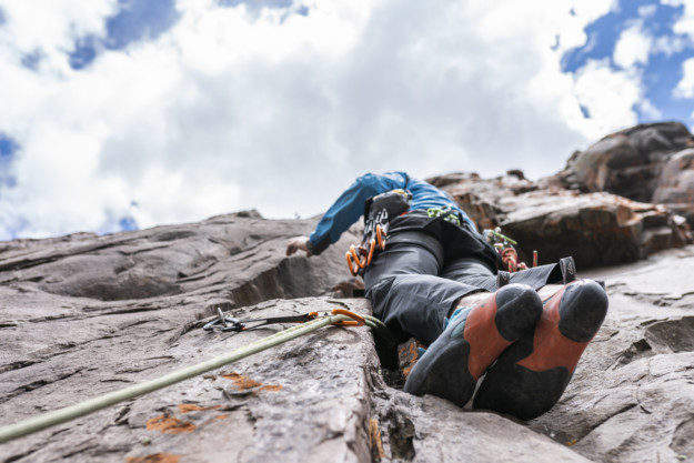 A climber in blue clothing ascends a steep rock face in Québec, focusing on a challenging route under a cloudy sky. The perspective emphasizes the height and the climber's grip on the rock.