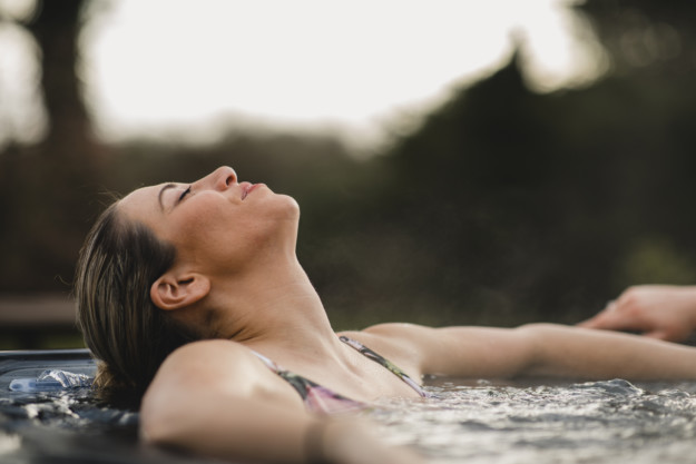 A woman relaxing in a hot tub outdoors in Québec, with her head tilted back, eyes closed, enjoying the serene environment.
