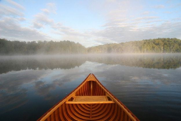 The front of a wooden canoe pointing towards a tranquil, mist-covered lake surrounded by forest under a clear sky at sunrise in Québec.