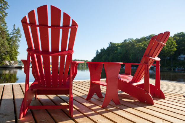 Two red adirondack chairs on a wooden dock facing a tranquil lake surrounded by forest in Québec, under a clear blue sky.