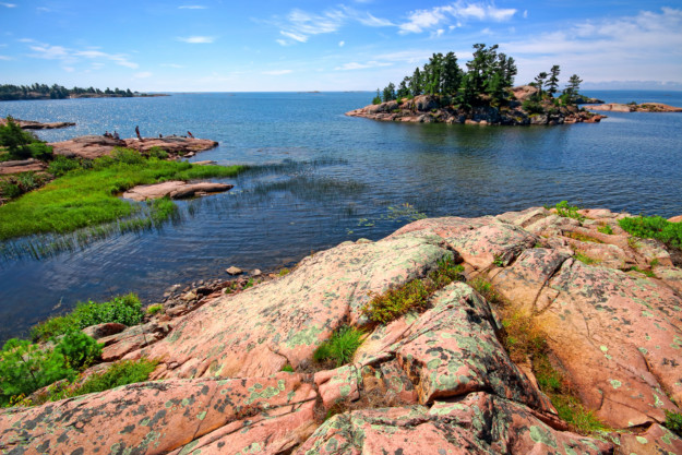A picturesque coastal scene in Québec featuring rugged pink granite rocks in the foreground, a serene blue lake, and small, lush green islands under a bright blue sky with scattered clouds.