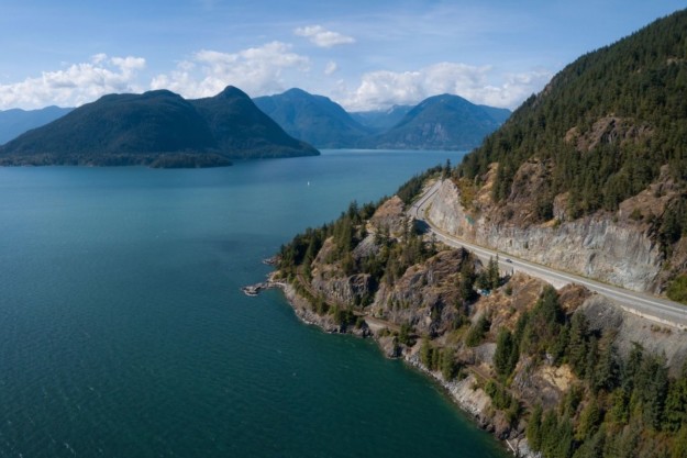 a coastal highway curves round a corner between the blue ocean and a hillside forest