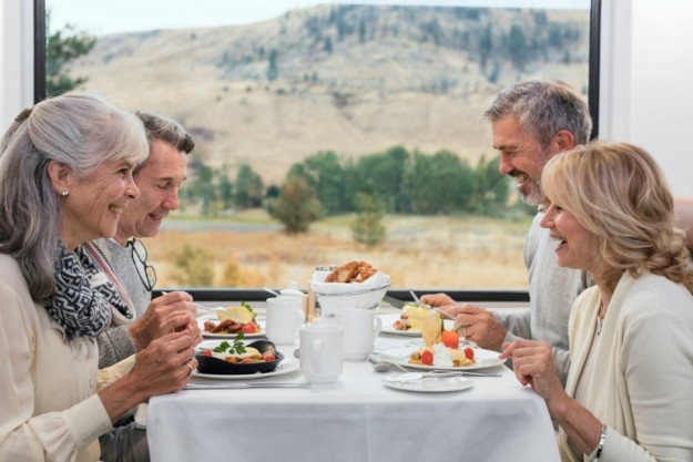 four people laugh and enjoy a meal aboard a train with a view of the mountains out of the window