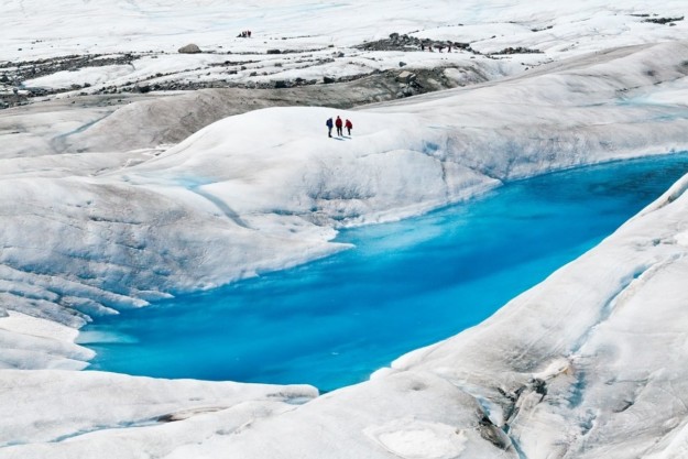 a turquoise blue pond sits in the middle of a white glacier with three people standing on the side