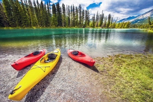 a yellow kayak flanked by two red kayaks sit on the edge of a lake of crystal clear water
