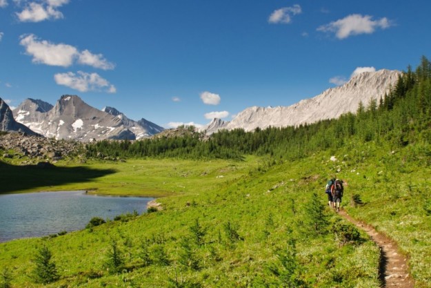 a pair of hikers walk along a trail with a lake on the left and a mountain in the background surrounded by greenery