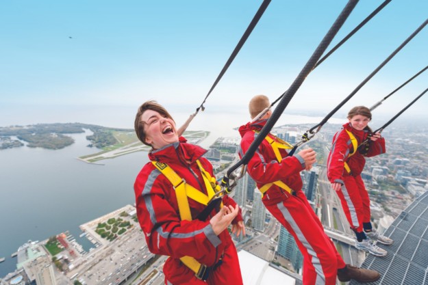three people in red jumpsuits hang over the edge at the CN tower