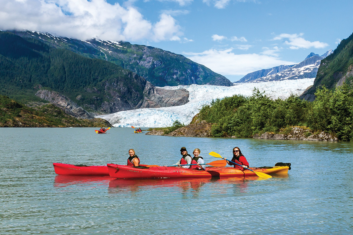 four smiling people sit in orange two man canoes with a glacier in the background