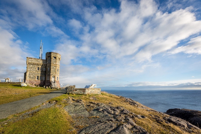 A scenic view of a historic stone tower on a rocky coastline in Nova Scotia under a wide sky with clouds, overlooking the ocean.