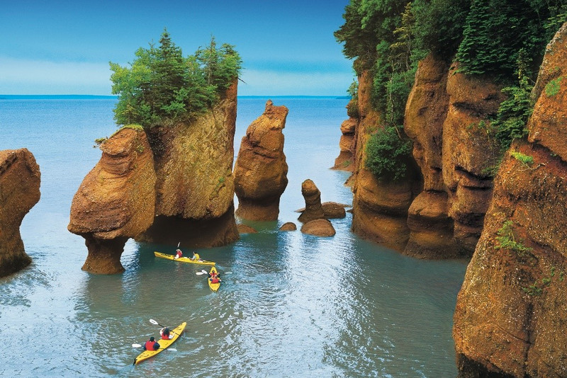 Three kayakers paddle near unique, tall rock formations and tree-topped islands in the calm, blue waters off the coast of Newfoundland and Labrador. The sky is clear and the surrounding vegetation is lush green