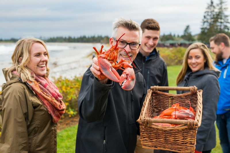 A joyful group of four adults on a beach in Nova Scotia, with a man in front holding up a large lobster towards the camera. A basket filled with lobsters is also visible in the foreground.
