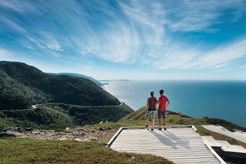 Two hikers with backpacks stand on a scenic viewpoint in Nova Scotia, overlooking a winding road and vast ocean with hills under a clear blue sky.