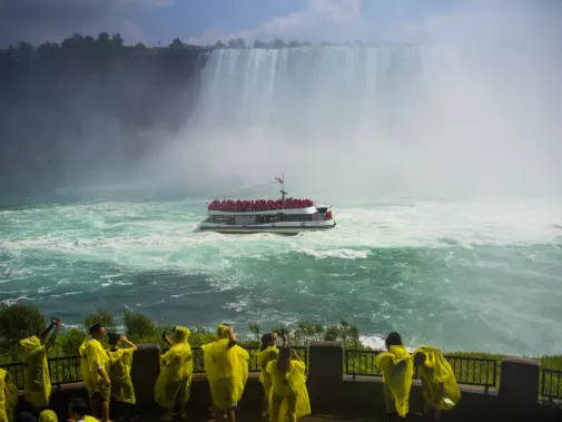 A tour boat approaches the misty base of Niagara Falls,viewed from a nearby platform where spectators in yellow raincoats watch