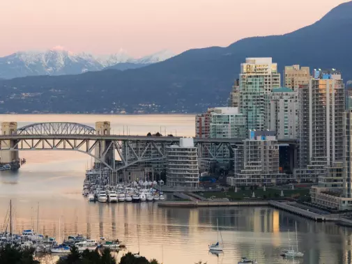 Twilight descends on the Vancouver waterfront, where modern high-rises meet calm waters,  and snowy mountain peaks in the distance adding a majestic backdrop.